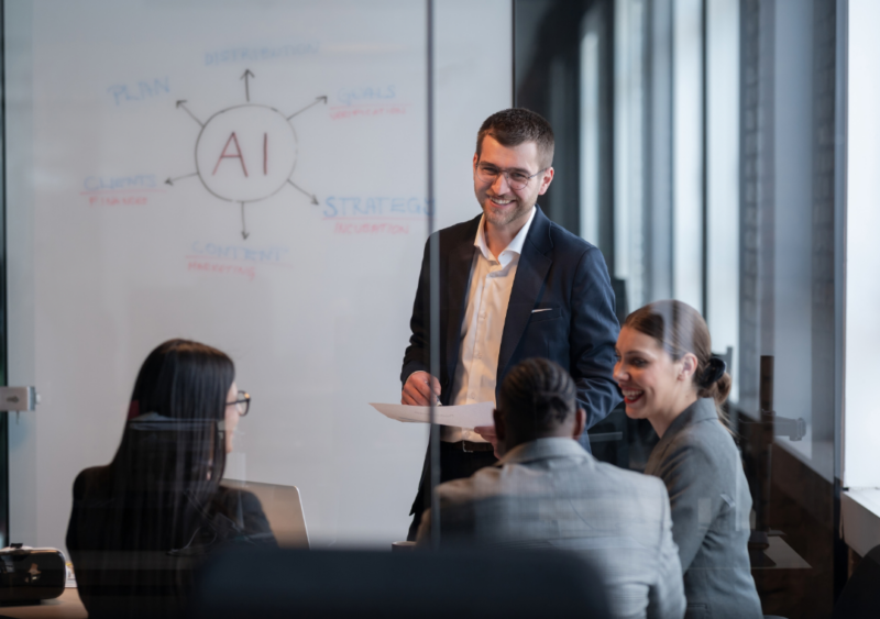 Man standing in meeting room, with three others. He is standing in-front of a whiteboard that says 'AI'. He is smiling.